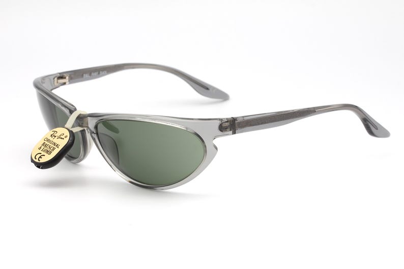 May include: Gray translucent sunglasses with green lenses. The sunglasses have a thin, curved frame and a small tag attached to the left arm that reads "Ray-Ban Original Bausch & Lomb CE".