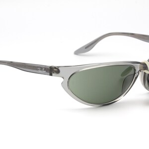 May include: Gray translucent sunglasses with green lenses. The sunglasses have a thin, curved frame and a Ray-Ban logo on the side. The sunglasses are attached to a small tag that reads "Original Ray-Ban Lenses".