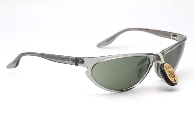 May include: Gray translucent sunglasses with green lenses. The sunglasses have a thin, curved frame and a Ray-Ban logo on the side. The sunglasses are attached to a small tag that reads "Original Ray-Ban Lenses".