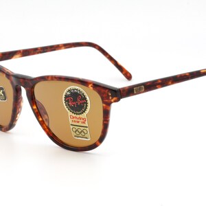 May include: Brown tortoiseshell sunglasses with brown lenses. The sunglasses have a classic rectangular frame shape. The lenses have a sticker that says "Ray-Ban Driving" and "Chromax". The sunglasses are on a white background.