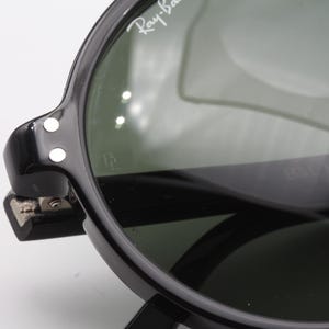 May include: Close-up of black Ray-Ban sunglasses with green lenses. The sunglasses have a metal hinge and two white dots on the side.