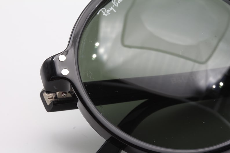 May include: Close-up of black Ray-Ban sunglasses with green lenses. The sunglasses have a metal hinge and two white dots on the side.