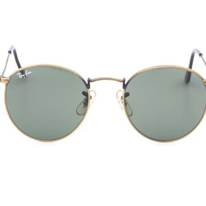 Puede incluir: Gafas de sol redondas con montura dorada y lentes verdes. El logotipo de Ray-Ban es visible en la lente derecha.