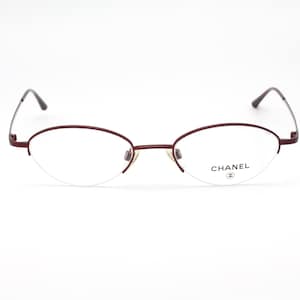 Puede incluir: Gafas Chanel con montura de metal burdeos y diseño semi-sin montura. Las lentes son transparentes y muestran el logo de Chanel. Las gafas tienen una forma ovalada clásica. La montura es de color rojo oscuro.
