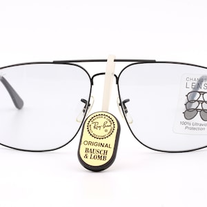 May include: Black square-shaped glasses with clear lenses. The glasses have a label on the right lens that says "CHANGEABLE LENSES" and "100% Ultraviolet Protection". A small tag with the words "ORIGINAL BAUSCH & LOMB" is hanging from the glasses.