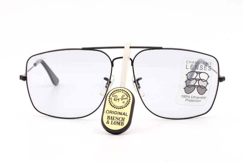 May include: Black square-shaped glasses with clear lenses. The glasses have a label on the right lens that says "CHANGEABLE LENSES" and "100% Ultraviolet Protection". A small tag with the words "ORIGINAL BAUSCH & LOMB" is hanging from the glasses.