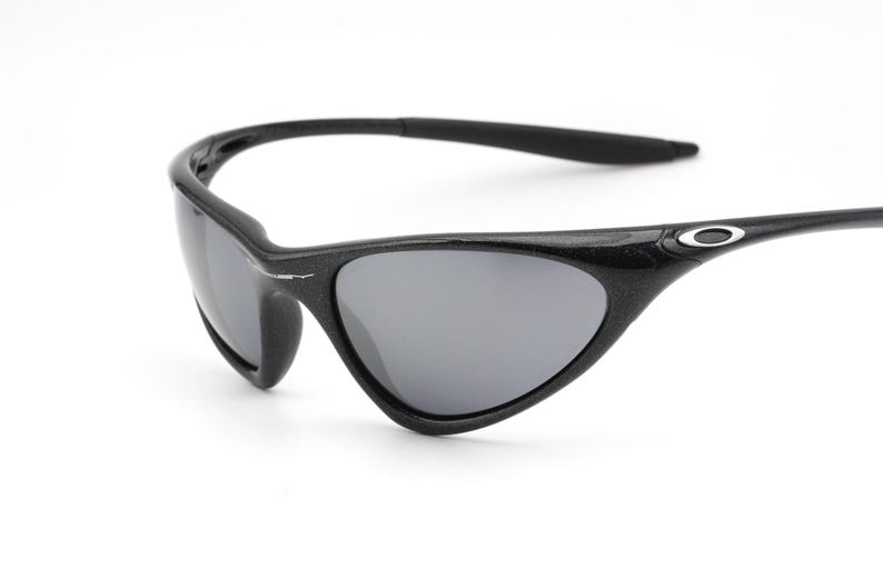 Puede incluir: Gafas de sol negras con lentes grises. Las gafas tienen una montura curvada y un peque&ntilde;o logotipo ovalado en la patilla. Las lentes son de color gris oscuro y la montura parece estar hecha de un material texturizado.