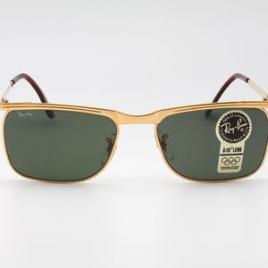 Op de afbeelding: Goudkleurige Ray-Ban zonnebril met groene glazen. De rechthoekige zonnebril heeft bruine pootjes. Een sticker op de lens leest "Ray-Ban" en "G-15 Lens".