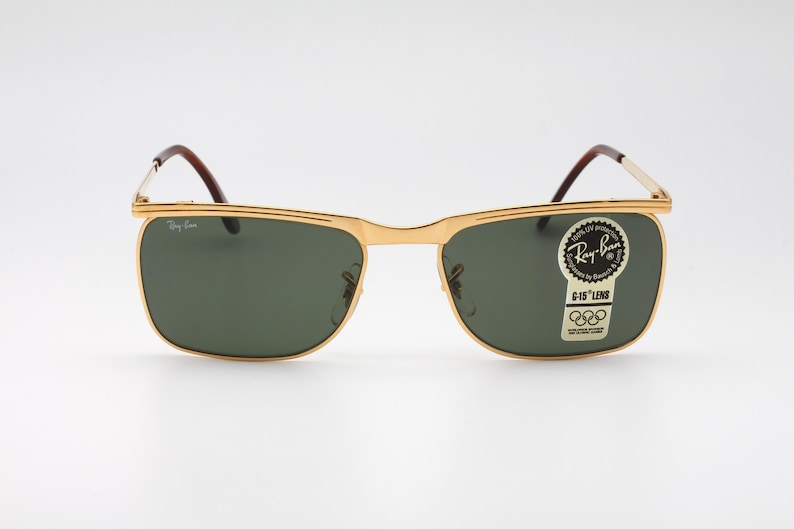 Op de afbeelding: Goudkleurige Ray-Ban zonnebril met groene glazen. De rechthoekige zonnebril heeft bruine pootjes. Een sticker op de lens leest "Ray-Ban" en "G-15 Lens".