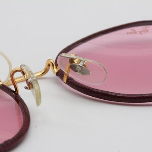 May include: A pair of pink round glasses with gold metal frames and burgundy leather accents. The glasses have a clear plastic nose bridge and a Ray-Ban logo on the right lens.