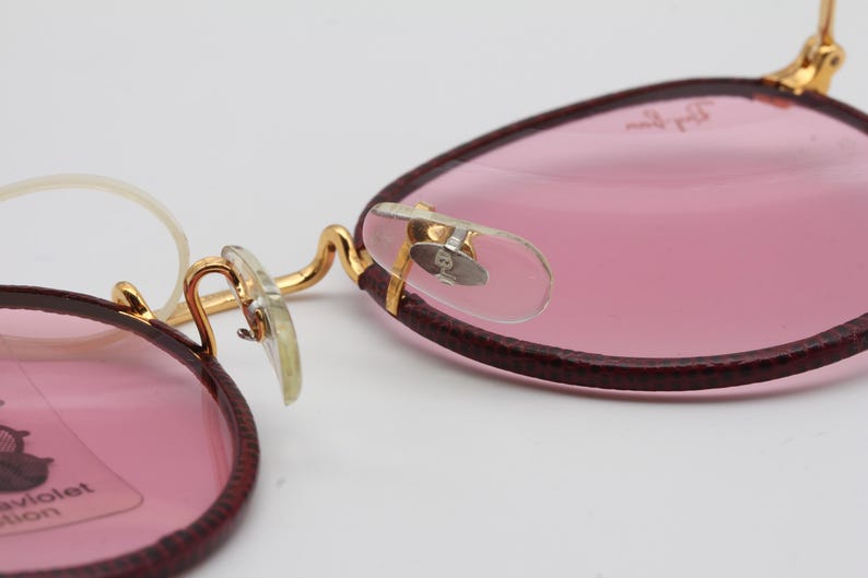 May include: A pair of pink round glasses with gold metal frames and burgundy leather accents. The glasses have a clear plastic nose bridge and a Ray-Ban logo on the right lens.