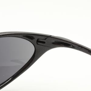 Puede incluir: Primer plano de gafas de sol negras con lentes grises oscuros. La montura es texturizada y tiene un dise&ntilde;o deportivo. Las gafas de sol est&aacute;n sobre un fondo blanco.