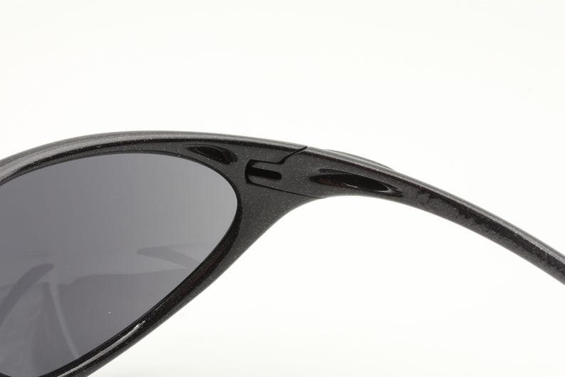 Puede incluir: Primer plano de gafas de sol negras con lentes grises oscuros. La montura es texturizada y tiene un dise&ntilde;o deportivo. Las gafas de sol est&aacute;n sobre un fondo blanco.