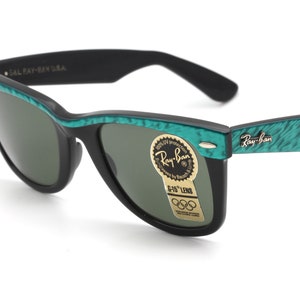 Puede incluir: Gafas de sol Ray-Ban negras con monturas de patr&oacute;n verde y negro. Las gafas tienen lentes verdes y una pegatina dorada que dice "Ray-Ban" y "G-15" Lens. La pegatina tambi&eacute;n tiene el logotipo de los anillos ol&iacute;mpicos.