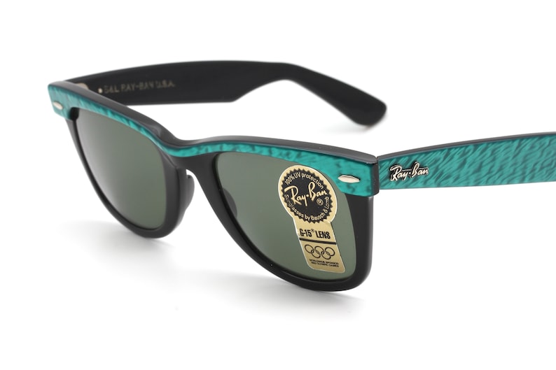 Puede incluir: Gafas de sol Ray-Ban negras con monturas de patr&oacute;n verde y negro. Las gafas tienen lentes verdes y una pegatina dorada que dice "Ray-Ban" y "G-15" Lens. La pegatina tambi&eacute;n tiene el logotipo de los anillos ol&iacute;mpicos.
