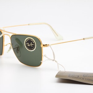 May include: Gold-tone aviator sunglasses with green lenses and a white Ray-Ban tag. The sunglasses have a white nose bridge and a white temple tip.