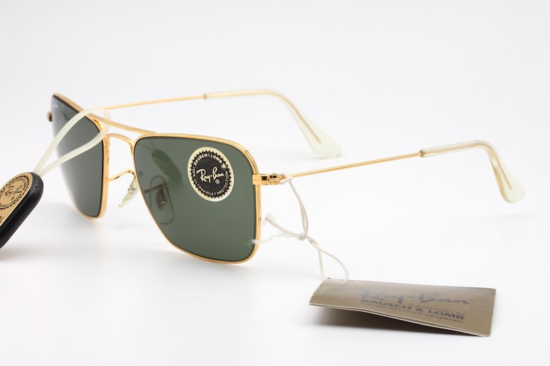 May include: Gold-tone aviator sunglasses with green lenses and a white Ray-Ban tag. The sunglasses have a white nose bridge and a white temple tip.