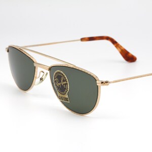May include: Gold-framed sunglasses with dark green lenses. The sunglasses have a double bridge and brown tortoiseshell-patterned temples. A Ray-Ban sticker is on the lens. The glasses are set against a white background.