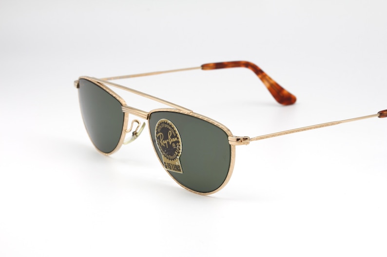 May include: Gold-framed sunglasses with dark green lenses. The sunglasses have a double bridge and brown tortoiseshell-patterned temples. A Ray-Ban sticker is on the lens. The glasses are set against a white background.