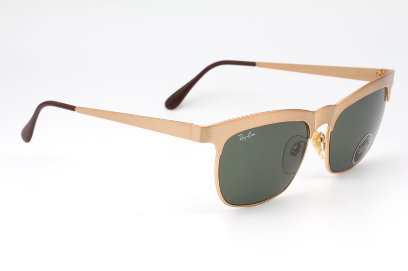 May include: Gold-tone Ray-Ban sunglasses with green lenses. The sunglasses have a classic aviator style with a thin metal frame.