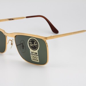 Op de afbeelding: Goudkleurige Ray-Ban zonnebril met donkergroene glazen. De zonnebril heeft een rechthoekige vorm en bruine pootjes. Op de lens zit een sticker met het Ray-Ban logo en Olympische ringen.