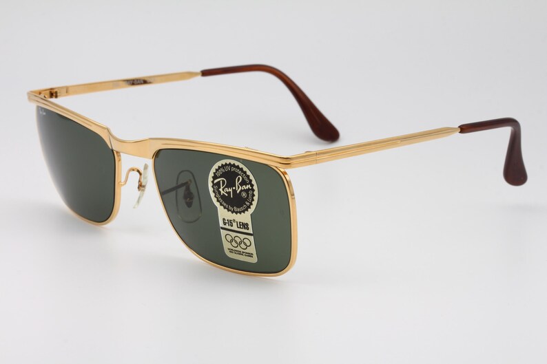 Op de afbeelding: Goudkleurige Ray-Ban zonnebril met donkergroene glazen. De zonnebril heeft een rechthoekige vorm en bruine pootjes. Op de lens zit een sticker met het Ray-Ban logo en Olympische ringen.