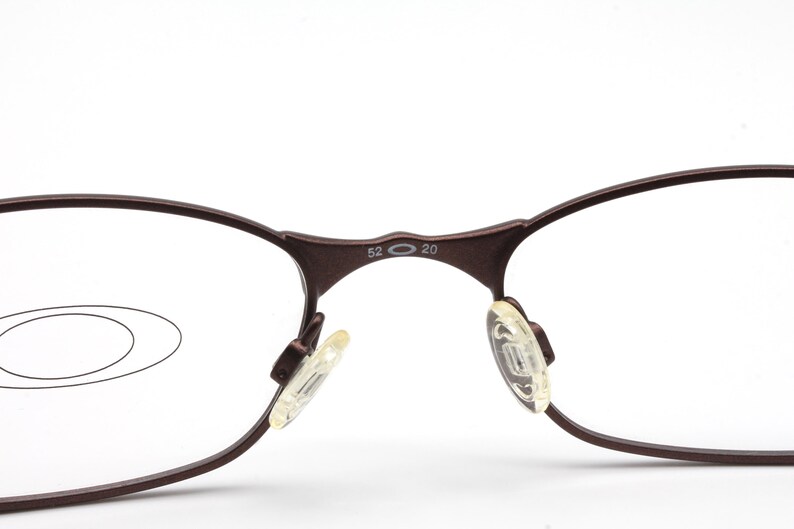 May include: Close-up of a pair of brown metal eyeglasses with clear lenses. The glasses have a unique oval shape and the arms are marked with "52 20".