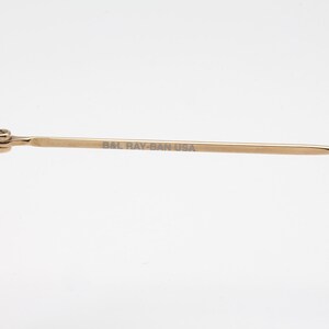 May include: Close-up of gold-toned sunglasses with dark lenses. The temple arm is visible, with the text "BAL RAY-BAN USA" printed on it. The sunglasses have a classic design.