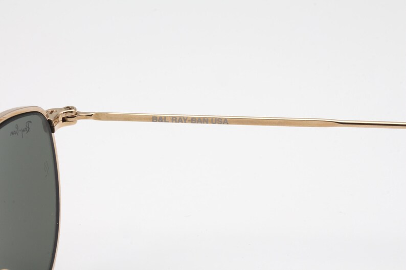 May include: Close-up of gold-toned sunglasses with dark lenses. The temple arm is visible, with the text "BAL RAY-BAN USA" printed on it. The sunglasses have a classic design.