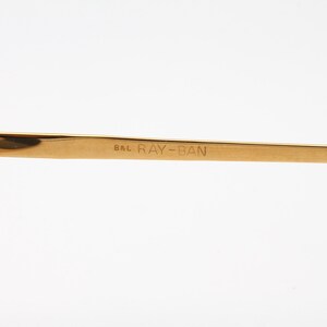 May include: Close-up of the gold-tone metal arm of a pair of Ray-Ban sunglasses. The arm is engraved with "B&L RAY-BAN".