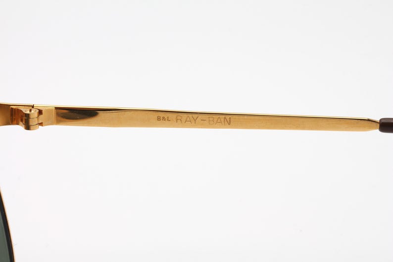 May include: Close-up of the gold-tone metal arm of a pair of Ray-Ban sunglasses. The arm is engraved with "B&L RAY-BAN".