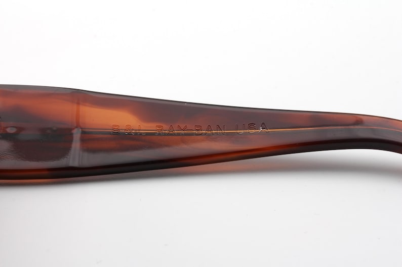 Puede incluir: Primer plano de una patilla de gafas de carey marr&oacute;n. El texto "B&L RAY-BAN USA" est&aacute; impreso en letras doradas a lo largo de la patilla. La patilla es curva y tiene un acabado brillante.
