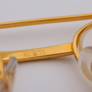 May include: A gold-colored metal frame of a pair of glasses with the inscription '© Cartier 60  □ 18' on the inside of the frame.
