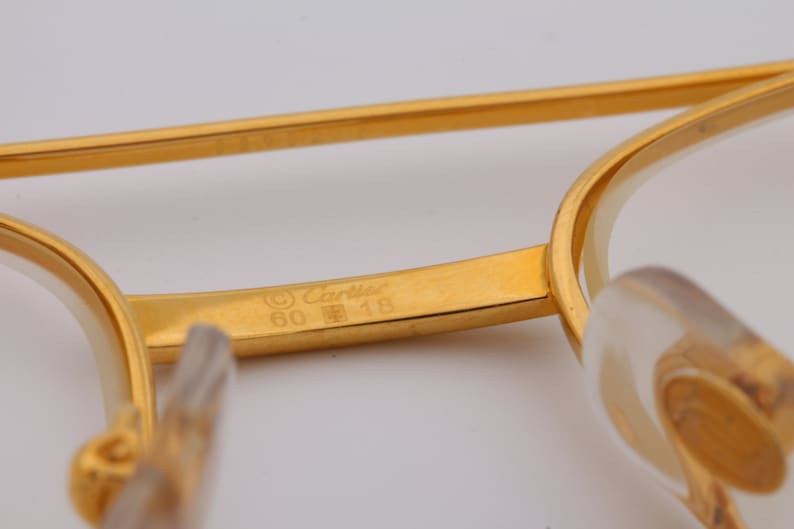 May include: A gold-colored metal frame of a pair of glasses with the inscription '© Cartier 60  □ 18' on the inside of the frame.