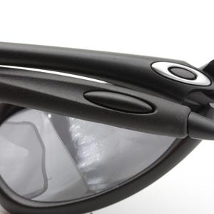 May include: Close-up of black sunglasses with gray lenses. The sunglasses have a sleek, modern design with a matte finish. The Oakley logo is visible on the temples. The frame is made of a durable material.