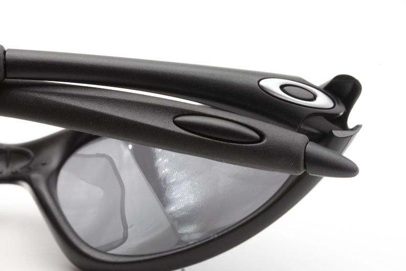 May include: Close-up of black sunglasses with gray lenses. The sunglasses have a sleek, modern design with a matte finish. The Oakley logo is visible on the temples. The frame is made of a durable material.