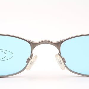 May include: Close-up of Oakley sunglasses with a silver frame and light blue lenses. The Oakley logo is visible on the left lens. The nose pads are clear. The glasses are set against a white background.