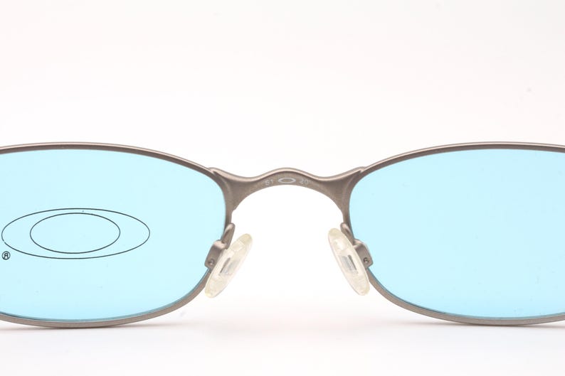 May include: Close-up of Oakley sunglasses with a silver frame and light blue lenses. The Oakley logo is visible on the left lens. The nose pads are clear. The glasses are set against a white background.