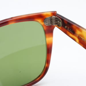 May include: Close-up of vintage sunglasses with a tortoiseshell frame and green lenses. The hinge is visible, and the arm of the glasses is marked with "B&L RAY-BAN USA".