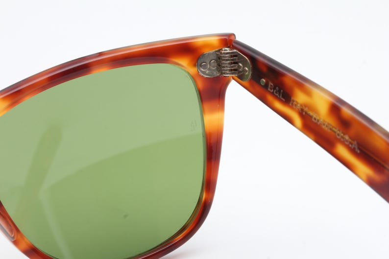 May include: Close-up of vintage sunglasses with a tortoiseshell frame and green lenses. The hinge is visible, and the arm of the glasses is marked with "B&L RAY-BAN USA".