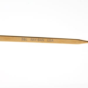 Op de afbeelding: Close-up van een goudkleurige brilpoot met de tekst "B&L RAY-BAN USA" in grijs gedrukt. De poot heeft een bruine punt. De bril is waarschijnlijk vintage of retro.