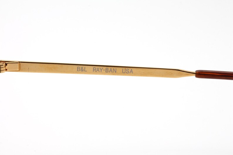 Op de afbeelding: Close-up van een goudkleurige brilpoot met de tekst "B&L RAY-BAN USA" in grijs gedrukt. De poot heeft een bruine punt. De bril is waarschijnlijk vintage of retro.