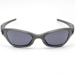 Może przedstawiać: Ciemnoszare okulary przeciwsłoneczne z zakrzywioną oprawką i ciemnymi soczewkami. Okulary przeciwsłoneczne mają sportowy design i są umieszczone na białym tle. Stylowy dodatek do aktywności na świeżym powietrzu.