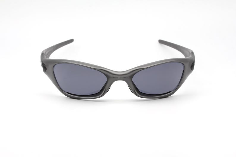 Może przedstawiać: Ciemnoszare okulary przeciwsłoneczne z zakrzywioną oprawką i ciemnymi soczewkami. Okulary przeciwsłoneczne mają sportowy design i są umieszczone na białym tle. Stylowy dodatek do aktywności na świeżym powietrzu.