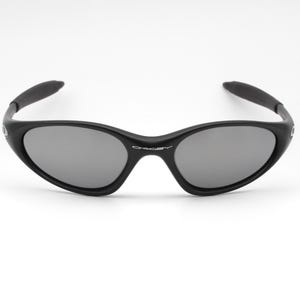 May include: Black sunglasses with gray lenses. The sunglasses have a curved frame and arms, and the brand name is visible on the bridge. The lenses are oval-shaped and reflect light. The sunglasses are set against a white background.