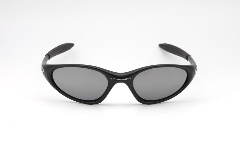 May include: Black sunglasses with gray lenses. The sunglasses have a curved frame and arms, and the brand name is visible on the bridge. The lenses are oval-shaped and reflect light. The sunglasses are set against a white background.