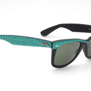 Puede incluir: Gafas de sol Ray-Ban Wayfarer negras con montura de patr&oacute;n verde azulado. Las gafas tienen lentes verdes y el logotipo de Ray-Ban es visible en la patilla izquierda.