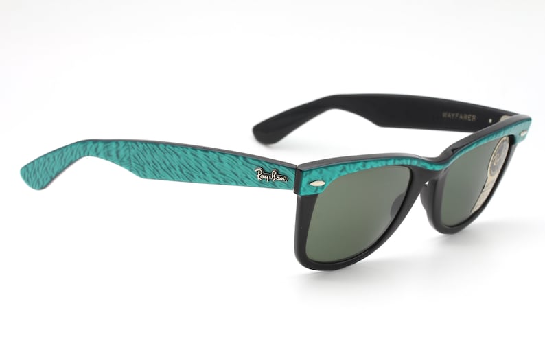 Puede incluir: Gafas de sol Ray-Ban Wayfarer negras con montura de patr&oacute;n verde azulado. Las gafas tienen lentes verdes y el logotipo de Ray-Ban es visible en la patilla izquierda.