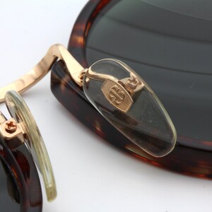 May include: Close-up of a pair of tortoiseshell sunglasses with gold accents. The sunglasses have a unique design with a hinge that allows the lenses to fold in. The brand name "B.B." is visible on the hinge.