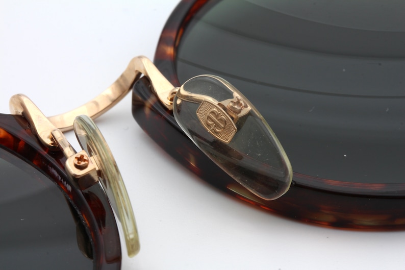 May include: Close-up of a pair of tortoiseshell sunglasses with gold accents. The sunglasses have a unique design with a hinge that allows the lenses to fold in. The brand name "B.B." is visible on the hinge.